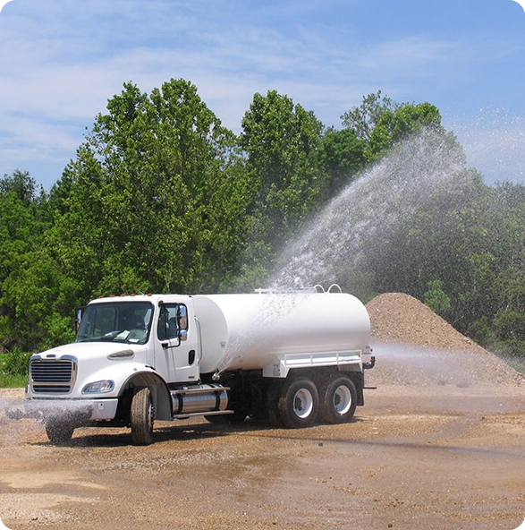 A Qathar Al Nada water truck sprays a large stream onto a dusty open area, surrounded by green trees under a clear blue sky, showcasing the company and information about its services.