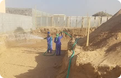 A Qathar Al Nada team in blue clothing works in a sandy, sunlit area at a water delivery project, surrounded by fencing and sand piles.