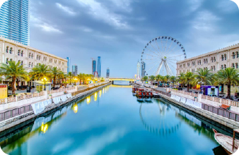 A scenic canal lined with greenery and modern buildings, leading towards a large Ferris wheel, with sweet water tanker supply nearby under a bright blue sky.