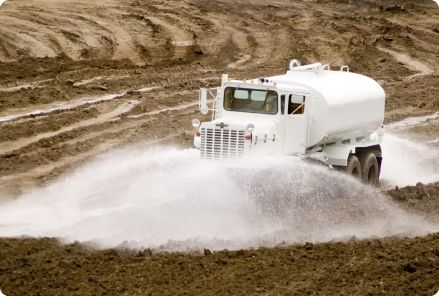A white truck sprays water from a water tank for construction on a muddy dirt track, creating a trail of mist for site maintenance and dust control.