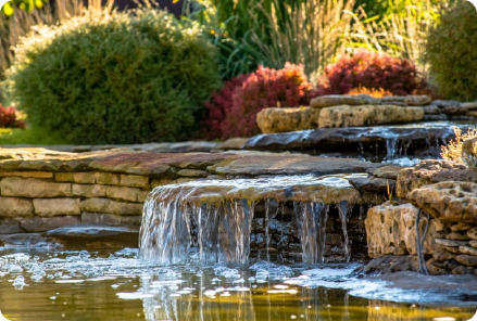 A tranquil garden scene with a small waterfall flowing over stone steps into a pond, surrounded by lush greenery and vibrant red bushes, with water supplied from a water tank for landscaping under soft sunlight.