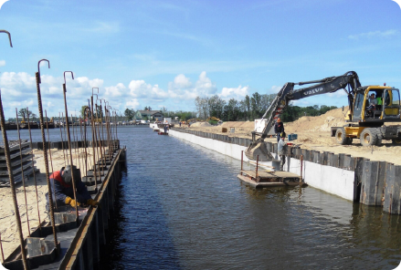 Construction site with a canal, featuring a crane placing concrete slabs, supported by water for piling sites, under clear skies for an efficient and active work environment.