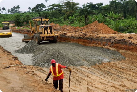 A construction worker in an orange vest directs machinery at a roadwork site, surrounded by red earth and green trees, with water for road construction being used on-site.