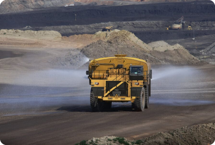 "A yellow construction truck sprays water for road construction on a dusty road at a mining site, with surrounding mounds of earth and a hazy sky creating an industrial scene.