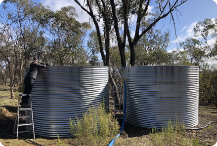 Two large, cylindrical metal tanks used for water for tank filling are split open, surrounded by dry grass and trees under a partly cloudy sky, suggesting a rural, isolated setting.
