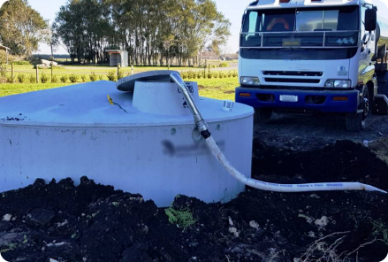 A large water tank used for water for tank filling with a hose connected to a truck in a rural setting. Trees and fields are visible in the background under bright daylight.