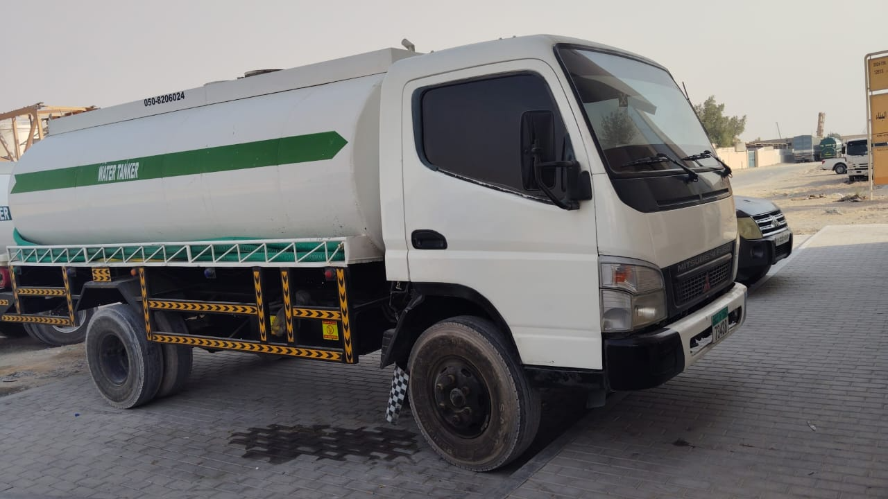 A white water tanker truck with a green stripe parked on a city street, part of sweet water tanker supply services, showing signs of wear on its tires.